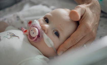 Toddler with nasal oxygen prongs insitu lying with head on pillow and sucking on dummy. Adult hand supporting head.